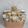 A gourmet snack and chocolate gift basket in a woven brown basket, wrapped in clear cellophane with a gold decorative bow, displayed on a wooden bench against a gray stone wall.