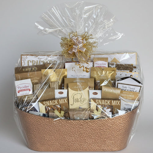 A gourmet snack and chocolate gift basket in a woven brown basket, wrapped in clear cellophane with a gold decorative bow, displayed on a wooden bench against a gray stone wall.