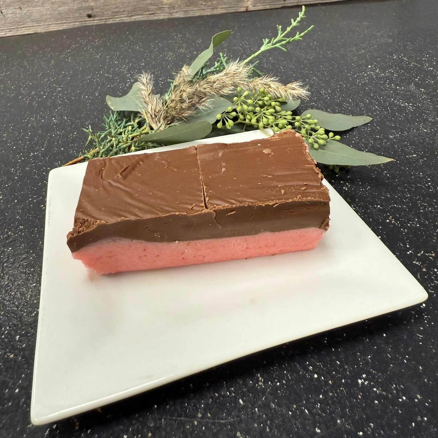Two-layered dessert bar with chocolate on top and pink interior on a white plate, set against a dark surface with greenery.